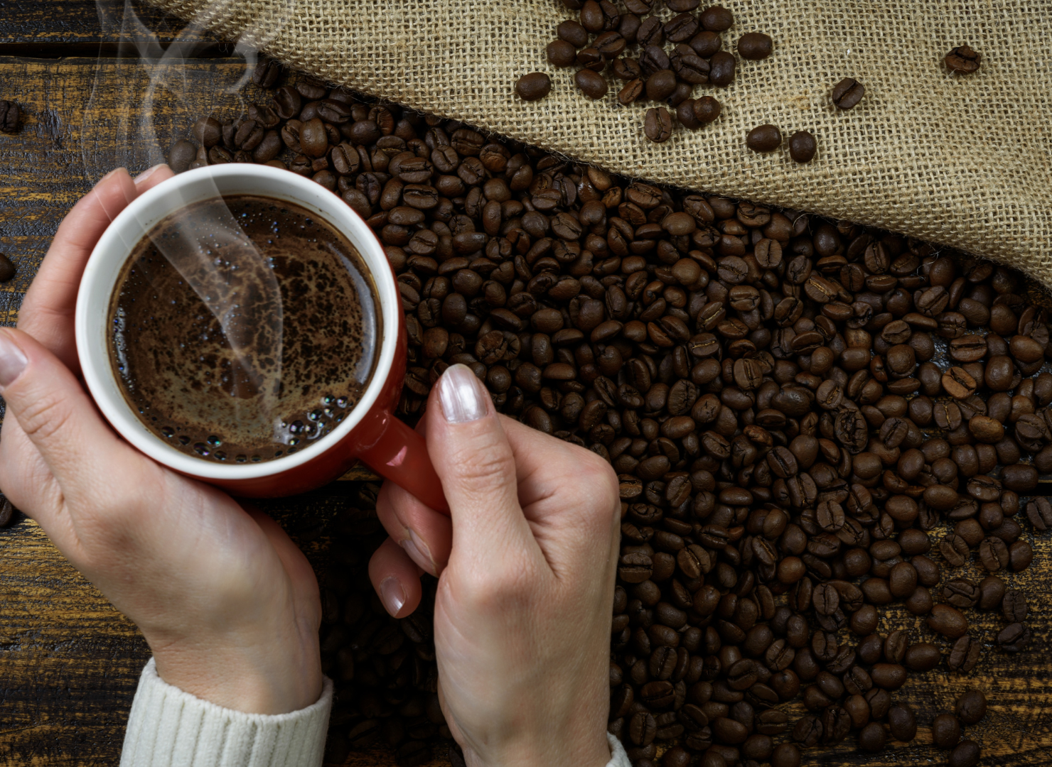 En hånd holder om en kop kaffe fyldt med rygende sort kaffe på et bord med kaffebønner liggende omkring.