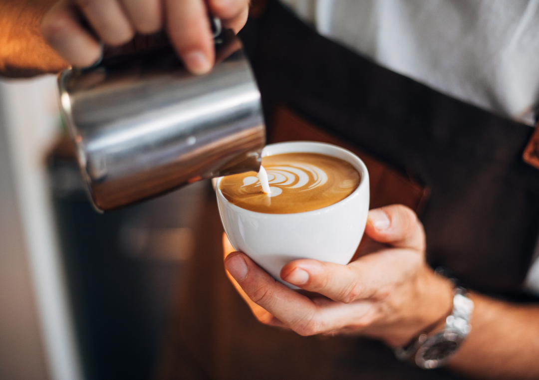 Mand hælder mælk fra en kande ned i en hvid kop kaffe.