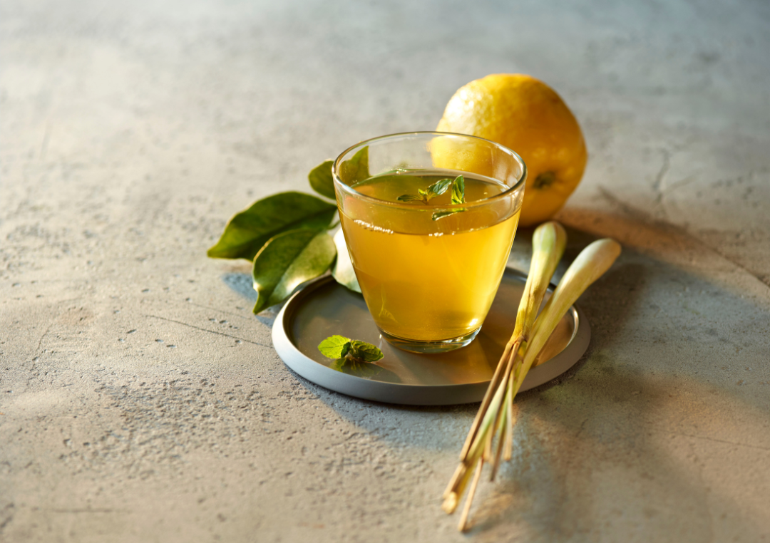 En lille glaskop med grøn te på en bordskånner med citron i baggrund og små blade bagved.