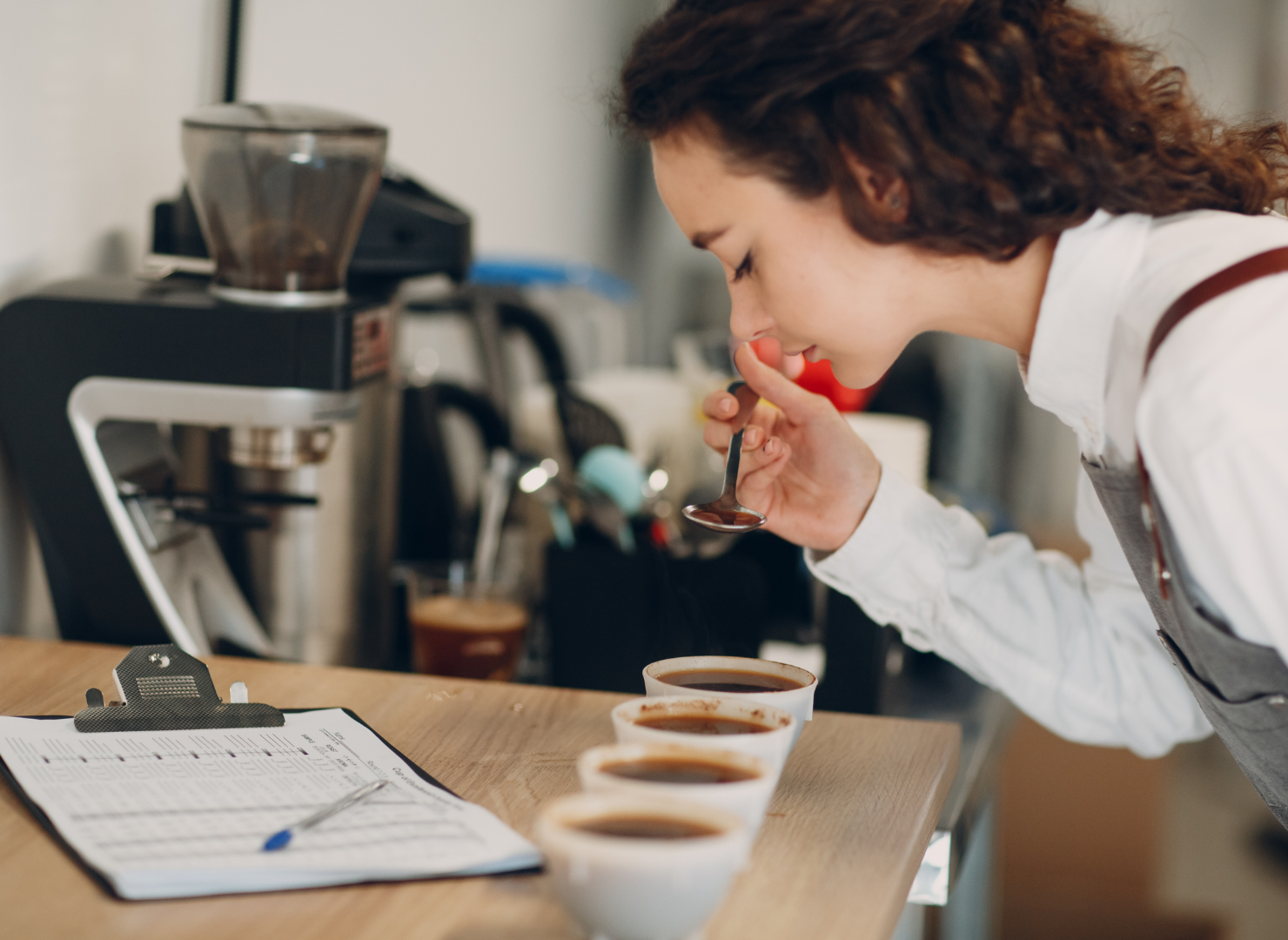 En kvinde står med en ske i hånden over 4 kopper sort kaffe.