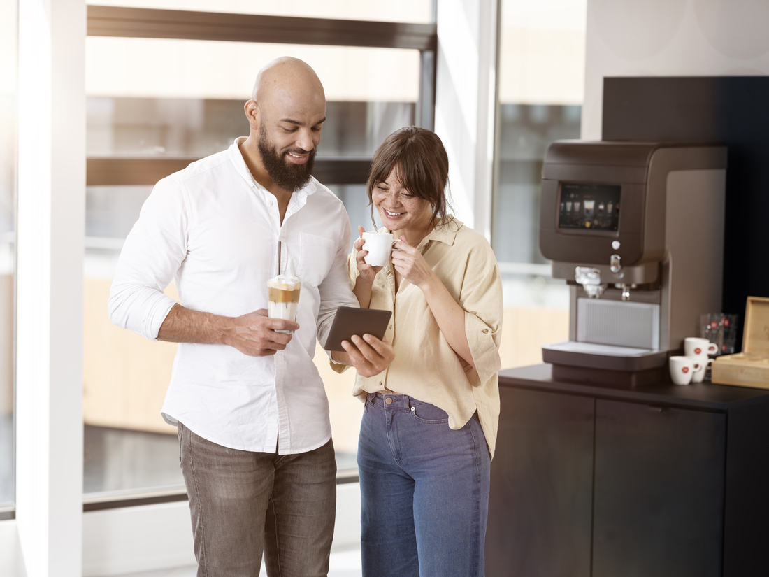 To personer ved kaffemaskine, den ene med tablet, begge med kaffe i moderne kontormiljø.