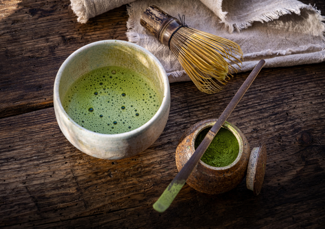 Et krus med matcha te i. Ved siden af en børste og en skål med grøn matchapulver.