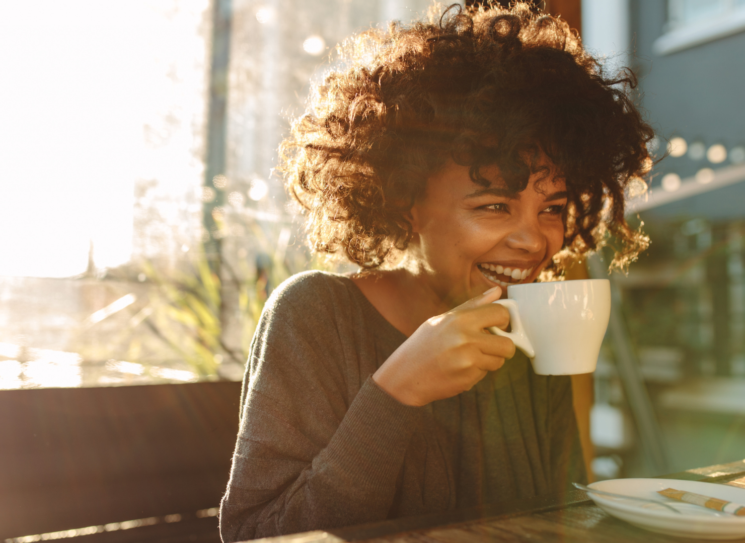 En kvinde med krøllet hår sidder med en kop kaffe og smiler