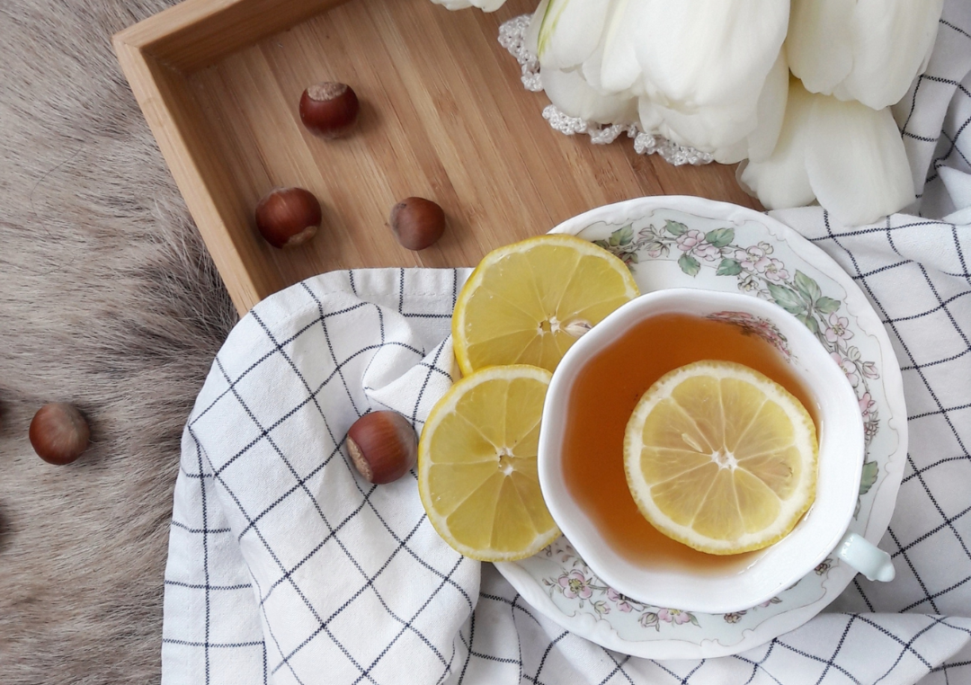 En kop med hvid te set oppe fra, på en bakke med små hasselnødder, citronskiver med et viskestykke.